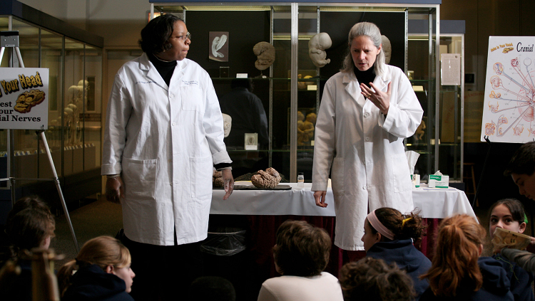 Two neuroscientists in white lab coats speaking to students during Brain Awareness Week.