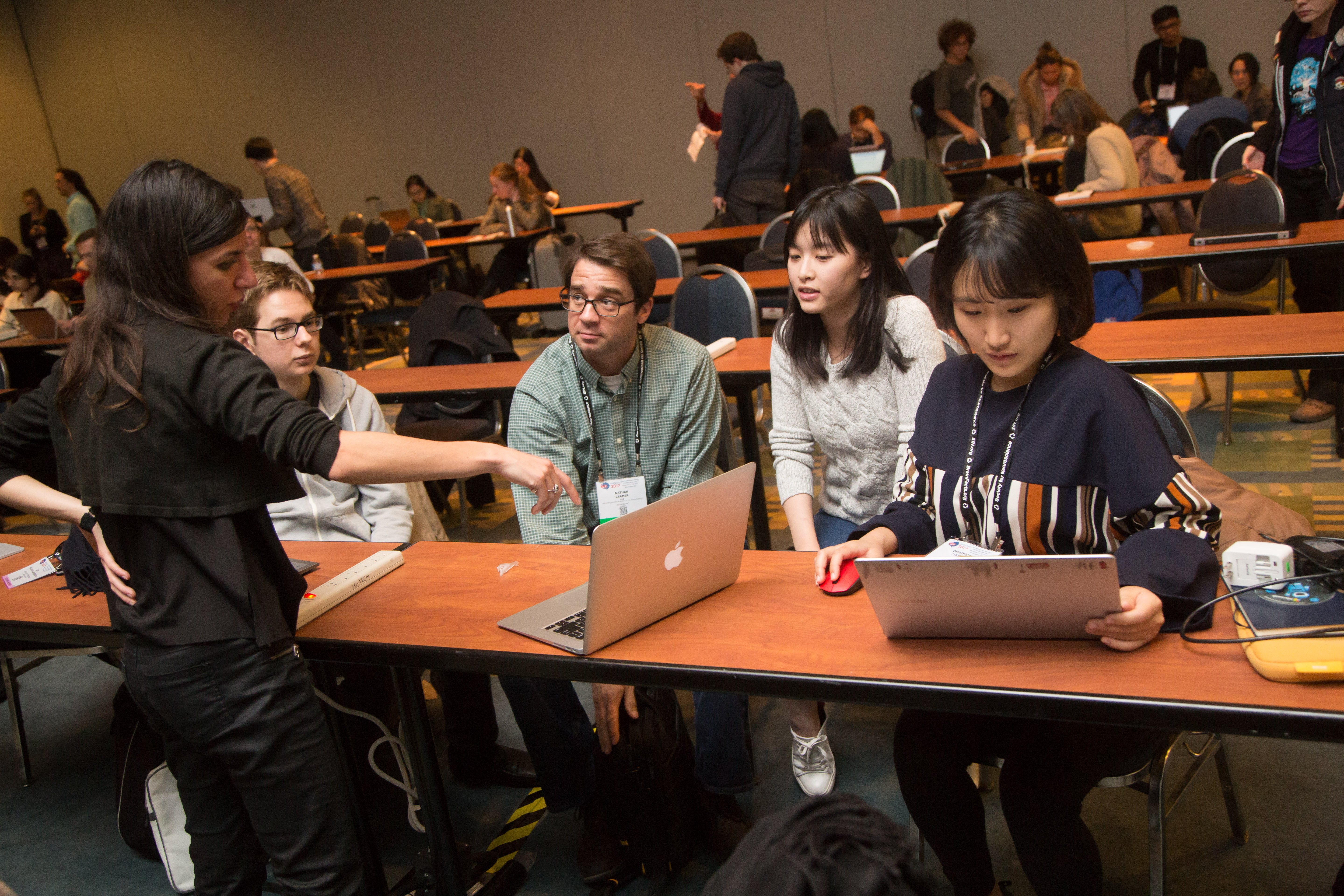 person pointing to a laptop screen while four people sitting at a table look