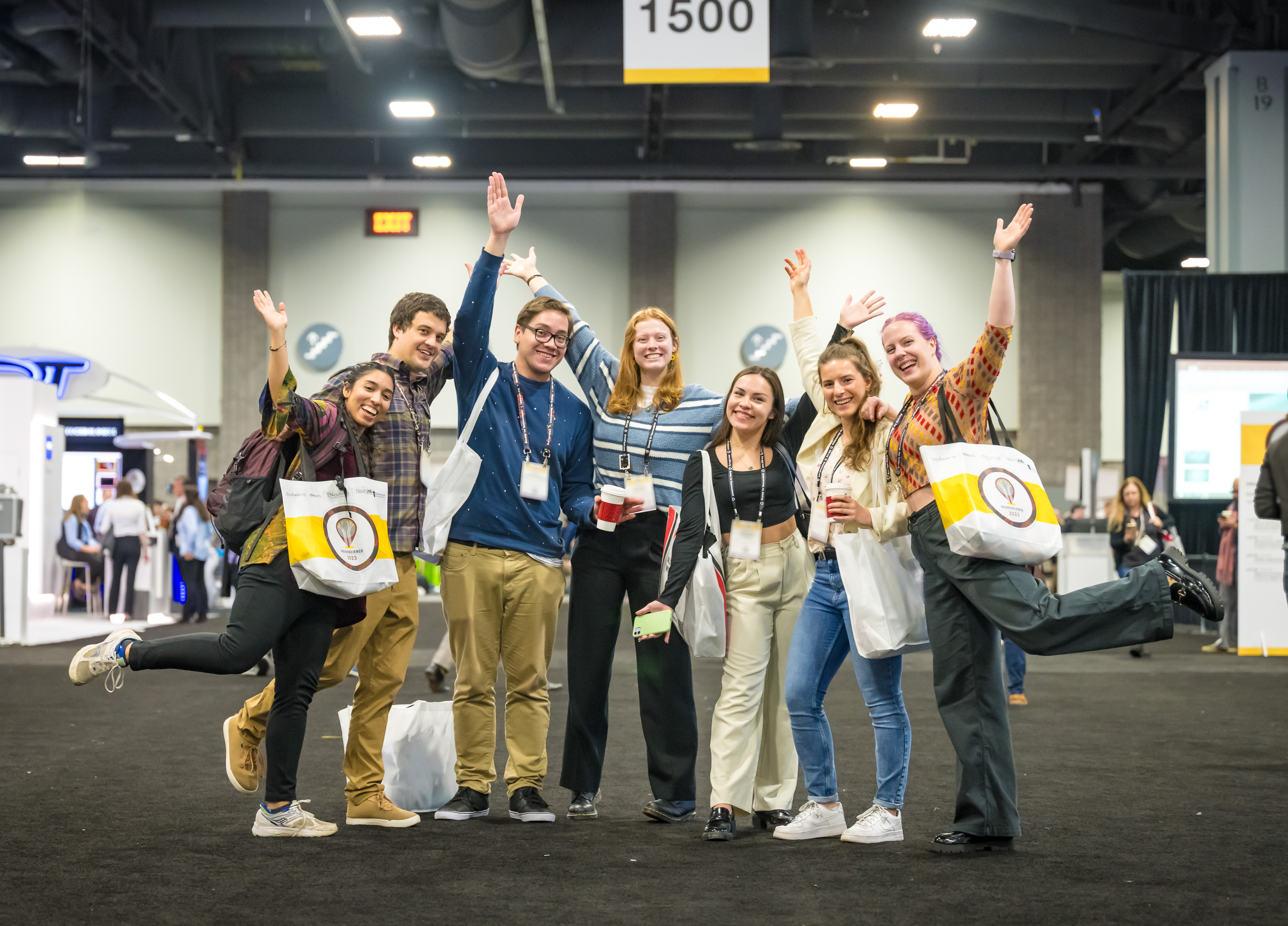 A group of seven attendees standing as a group in the exhibit hall raising their arms and smiling